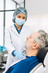 Female dentist wearing medical mask and cap looking at male patient in her office