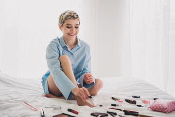 Smiling young woman in blue shirt relaxing indoors surrounded by cosmetics on bed in bright bedroom...