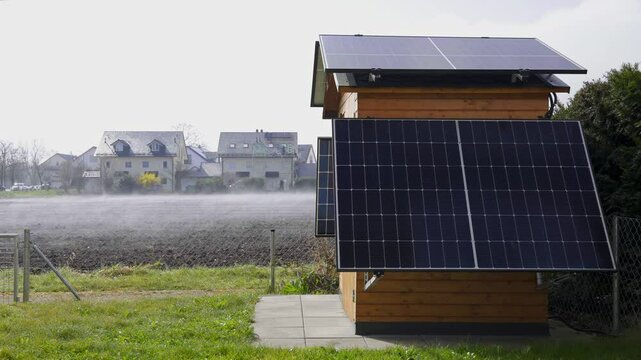 Balkonkraftwerk mit Nebel der &uuml;ber das Feld im Hintergrund zieht. An einem Hausdach im Hintergrund wird gerade eine PV Anlage installiert.