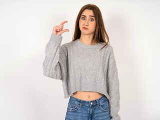 Caucasian young girl wearing grey sweater purses lip and gestures with hand, shows something very little.