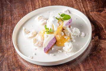  A slice of purple mousse cake topped with white meringue and vibrant mango sauce, garnished with mint leaves, elegantly served on a round white plate against a wooden background.