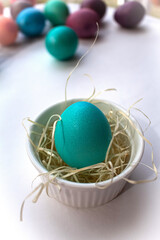 A green and blue Easter egg stands in a white porcelain tartlet with straw on a white background