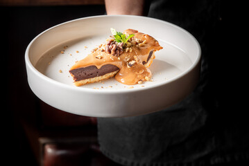 A server holds a slice of rich chocolate tart drizzled with caramel sauce and topped with crushed nuts and a mint leaf, elegantly served on a round white plate against a dark background.