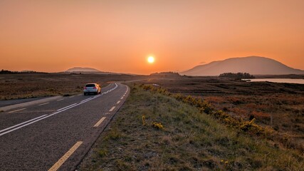 Beautiful landscape scenery with white car driving on empty scenic road trough nature and mountains at sunset, Connemara national park, Galway Ireland	
