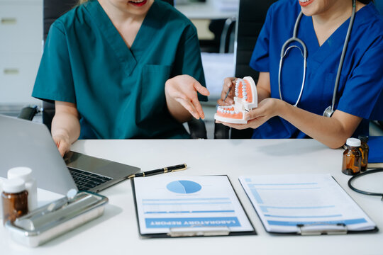Dentist sitting at table with jaw samples tooth model and working with tablet and laptop in dental modern office - Powered by Adobe