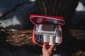 A person is standing in front of a large tree while holding a bright red first aid kit, which is...