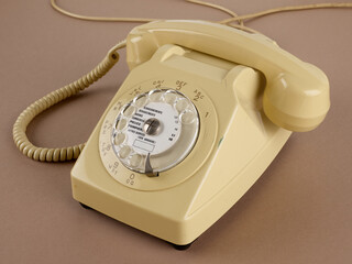 Un téléphone à cadran rotatif de couleur beige, avec écouteur secondaire, avec ses câbles. Des chiffres et des lettres pour la numérotation sont visibles. Sur fond marron.