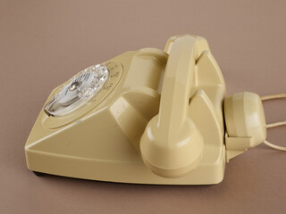 Un téléphone de profil à cadran rotatif de couleur beige, avec écouteur secondaire, avec ses câbles. Des chiffres et des lettres pour la numérotation sont visibles. Sur fond marron.