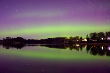 Northern lights glow green and purple over a calm lake at night, reflecting stars and trees in the water.