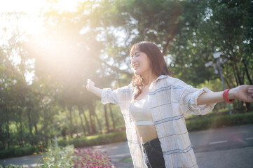 Asian woman enjoying summer, breathing fresh air with arms outstretched in sunny park, surrounded...