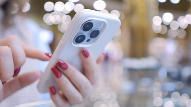 Woman is holding a white iPhone with a pink manicured hand. She is looking at the screen and she is texting