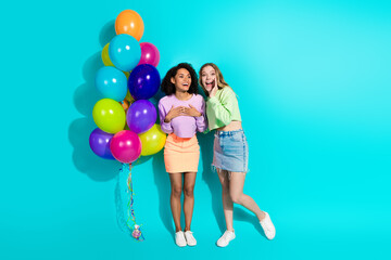 Fototapeta premium Two joyful young women with colorful balloons, standing against a vibrant teal background