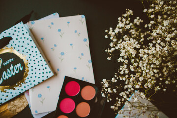 Eyeshadow palette, feathers, makeup brushes, flowers, notebooks and holiday bag on black paper background