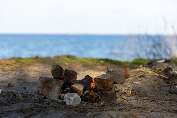 Obraz premium A Serene Lakeside Campsite Featuring a Cozy Campfire Surrounded by Rocks Against a Backdrop of Calm Water and Lush Greenery Beneath a Clear Blue Sky