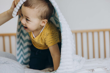 Smiling toddler having fun on bed with blanket. A baby playing hide and seek or peek a boo.