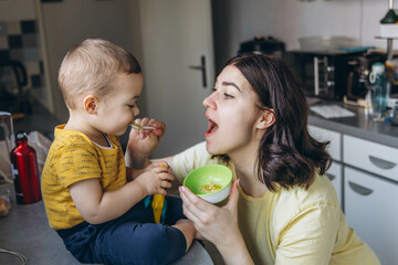 Fototapeta premium Mother feeding her son with a spoon in the kitchen, sharing a joyful and loving moment of motherhood