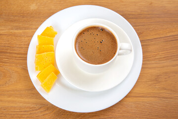 top-down view of a white cup of espresso with a frothy surface on a saucer, paired with slices of yellow cheese on a wooden table. Minimalistic and rustic setting.