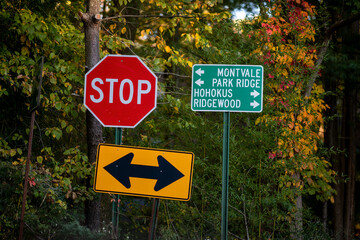 At a road intersection, a stop sign and directional signs point towards Montvale, Park Ridge, Hohokus, and Ridgewood amidst vibrant autumn leaves. Sunlight filters through the trees.