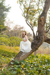 A girl in white dress sits on a tree holding flowers and feels the sunshine