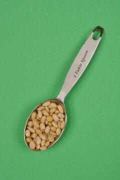 A single tablespoon of pine nuts in a metal spoon labeled - 1 Table Spoon - shot from above on a green background