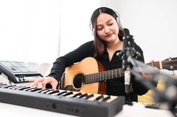 Beautiful Asian teen girl playing guitar, composing music in home recording studio using computer,...