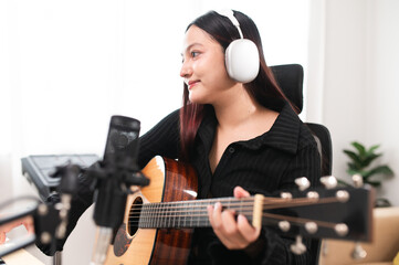 Beautiful Asian teen girl playing guitar, composing music in home recording studio using computer,...