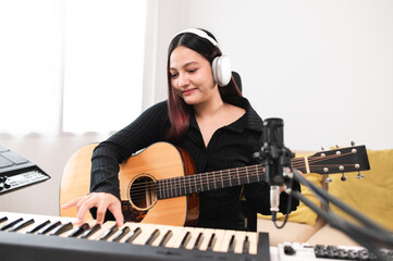 Beautiful Asian teen girl playing guitar, composing music in home recording studio using computer, headphones, condenser microphone, tablet and keyboard for mixing and mastering music.
