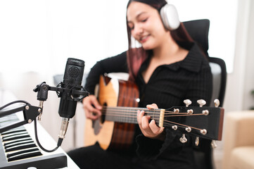 Beautiful Asian teen girl playing guitar, composing music in home recording studio using computer,...