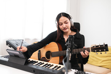 Beautiful Asian teen girl playing guitar, composing music in home recording studio using computer,...