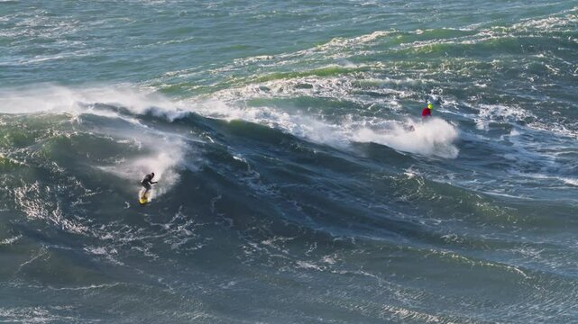 Surfer is riding big wave he is accompanied by a jet ski nearby. Extremal big wave surfing. Nazare, Portugal