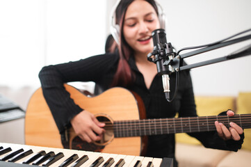 Beautiful Asian teen girl playing guitar, composing music in home recording studio using computer,...