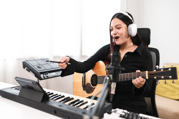 Beautiful Asian teen girl playing guitar, composing music in home recording studio using computer,...