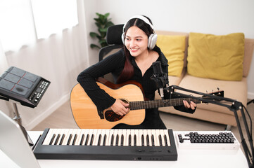 Beautiful Asian teen girl playing guitar, composing music in home recording studio using computer,...