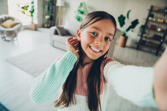 Smiling young girl in casual knitwear taking a selfie in a bright and cozy living room interior, enjoying a relaxed day at home. - Powered by Adobe