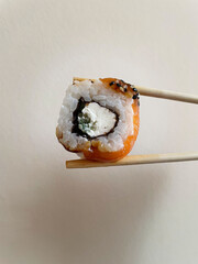 Small square piece of roll with rice, cheese, cucumber, nori and salmon is clamped between two wooden light brown chopsticks on beige background, close-up, side view. Japanese cuisine.