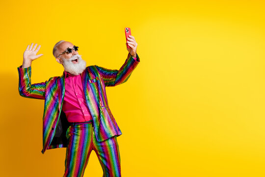 Photo of extravagant funny aged man smart phone selfie wave hi empty space wear shiny costume isolated on yellow color background
