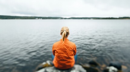 Fototapeta premium A person in an orange jacket gazes at the calm water under a cloudy sky, enjoying a peaceful moment alone