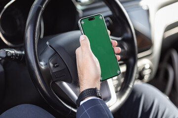 businessman holds smartphone green screen design space in one hand on the steering wheel. He is in car parked or in traffic jam in an urban environment. Concept: using phone technology