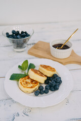 A plate of homemade cheese pancakes served with fresh blueberries. Accompanied by syrup and a mint garnish, this breakfast inspires a feeling of freshness and culinary delight.