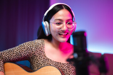 Asian teen girl playing guitar, composing music in home recording studio, LED lighting, at night using computer, headphones, condenser microphone, tablet and keyboard for mixing and mastering music.