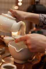 Hands pouring steaming tea from a white pot into a delicate cup in a warm atmosphere. Soft lights and wooden surfaces create a tranquil and cozy tea experience. Tea ceremony. Japanese tea party
