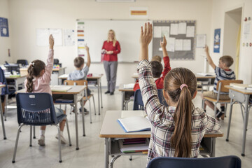 A bright classroom scene with students raising their hands enthusiastically The classroom has white walls and fluorescent lighting Students are seated at light