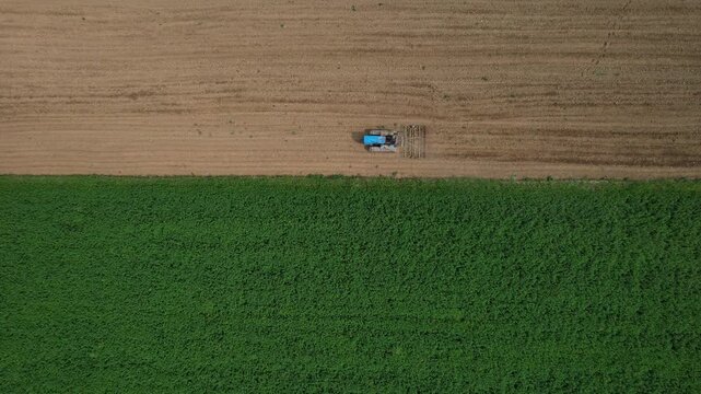 Slow Aerial view capturing blue tractor plowing agricultural landscape, creating striking geometric pattern of tilled brown soil contrasting with vibrant green crop rows