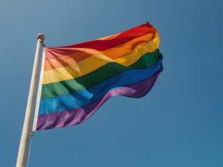 Rainbow Flag in the Wind: A vibrant rainbow flag waves proudly against a backdrop of a clear, brilliant blue sky, symbolizing hope, diversity, and acceptance. 