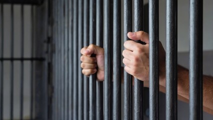 A prisoner holds onto the bars of a jail cell, emphasizing confinement and hopelessness