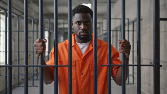 A prisoner wearing an orange shirt stands behind bars, conveying a sense of confinement and imprisonment