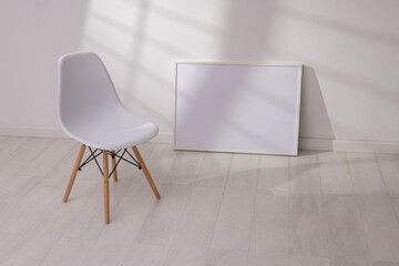 Mockup of empty frame in modern minimalist interior with chair and morning light on white wall background. Template for artwork, painting, photo or poster