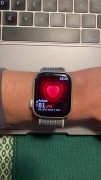 Mu&ntilde;eca del brazo izquierdo con reloj digital mostrando un video de los latidos por minuto LPM, se ve una imagen de coraz&oacute;n latiendo, apoyado en una mesa cerca de un teclado, vista vertical
