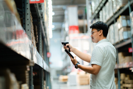 A man is standing in a warehouse, holding a barcode scanner and looking at a box. He is focused on scanning the barcode, possibly to check the inventory or to complete a task