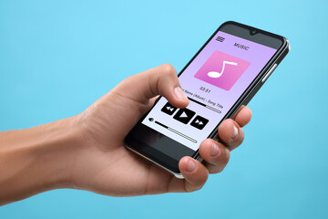 Man using mobile phone to listen to music on light blue background, closeup. Open music player app on device screen